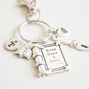 May include: Silver-coloured keychain with various charms. Charms include a book, a scroll with "Once Upon A Time" text, a pen nib, a heart-shaped key, and a letter "F". The keychain is attached to a silver clasp.