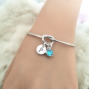 May include: Silver knot bangle bracelet with a charm featuring a letter 'P' and a turquoise blue crystal.