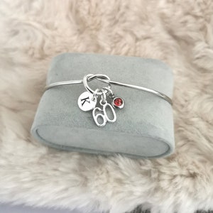 K&ouml;nnte beinhalten: Silbernes Knoten-Armband mit Charms. Die Charms beinhalten einen Buchstaben 'K', die Zahl '60' und einen roten Edelstein.