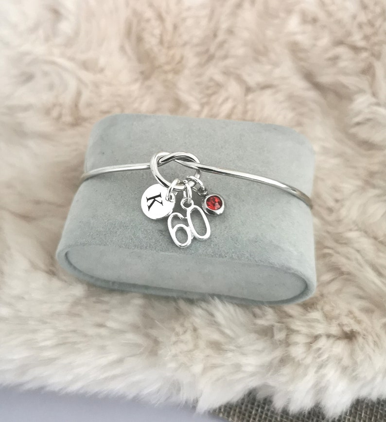 K&ouml;nnte beinhalten: Silbernes Knoten-Armband mit Charms. Die Charms beinhalten einen Buchstaben 'K', die Zahl '60' und einen roten Edelstein.