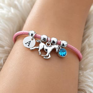 May include: A pink cord bracelet featuring a silver-toned horse charm, a circular silver-toned charm engraved with the letter A, three silver-toned beads, and a small round bright blue crystal charm attached to the cord with silver-toned hardware.