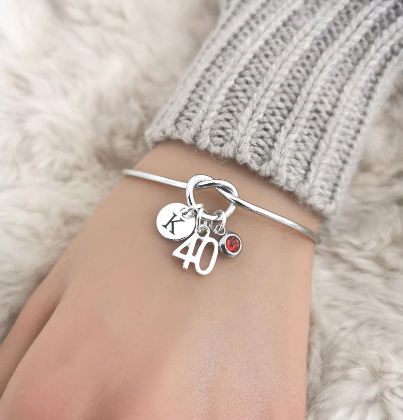 May include: A silver bangle bracelet with a knot design. It has charms including the number "40", a red gemstone, and a disc with the letter "K". The bracelet is worn on a wrist.