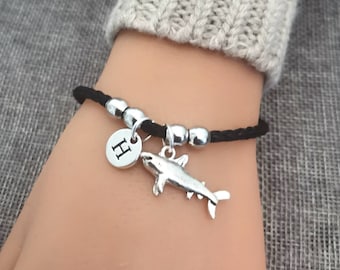 Könnte beinhalten: Schwarzes Kordelarmband mit silbernen Perlen und einem silbernen Hai-Anhänger. Der Anhänger hat eine kleine silberne Scheibe mit dem Buchstaben "H" darauf.