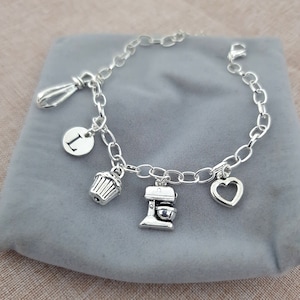 May include: A silver charm bracelet with a variety of charms, including a letter "L", a cupcake, a mixer, and a heart. The bracelet is on a gray background.