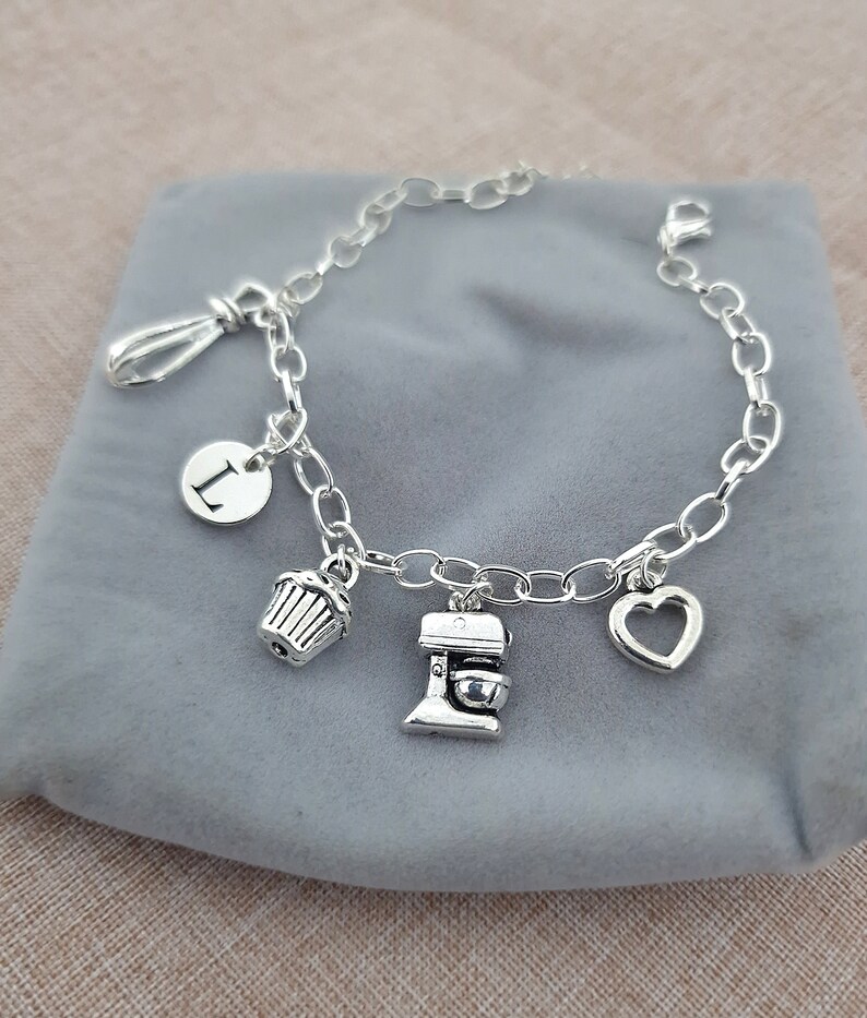May include: A silver charm bracelet with a variety of charms, including a letter "L", a cupcake, a mixer, and a heart. The bracelet is on a gray background.