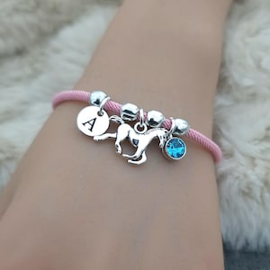 K&ouml;nnte beinhalten: Rosafarbenes Kordelarmband mit silbernen Perlen, einem silbernen Pferdeanh&auml;nger, einem silbernen Anh&auml;nger mit dem Buchstaben A und einem hellblauen Kristallanh&auml;nger.
