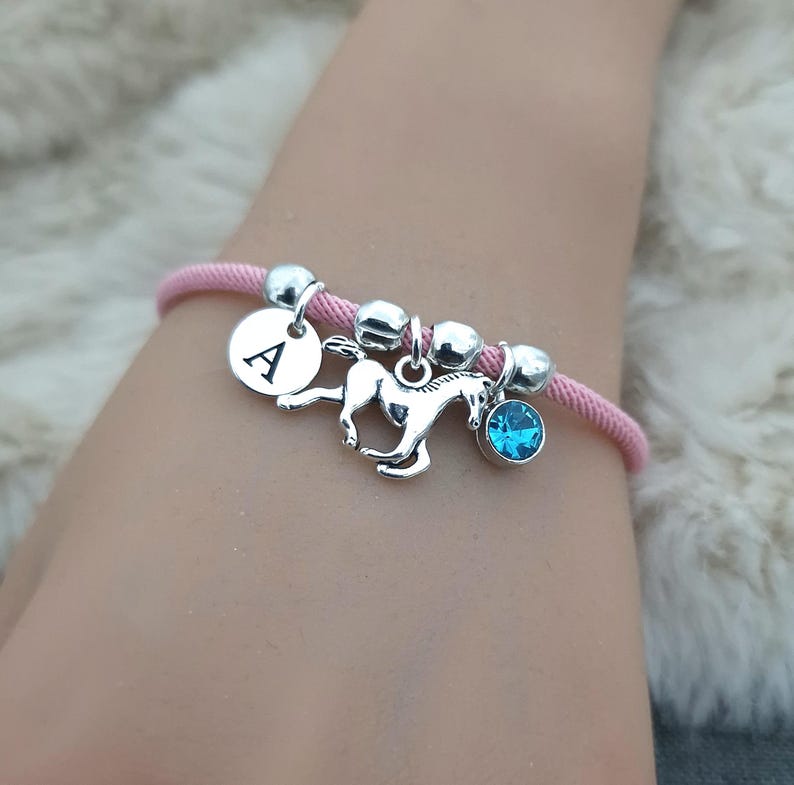 K&ouml;nnte beinhalten: Rosafarbenes Kordelarmband mit silbernen Perlen, einem silbernen Pferdeanh&auml;nger, einem silbernen Anh&auml;nger mit dem Buchstaben A und einem hellblauen Kristallanh&auml;nger.