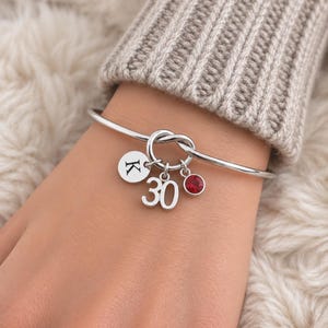 Op de afbeelding: Een zilverkleurige armband met een knoopontwerp in het midden. Drie bedels hangen aan de knoop: een ronde schijf gegraveerd met de letter K, het getal 30 in een vetgedrukt lettertype en een kleine, ronde, gefacetteerde rode kristal.