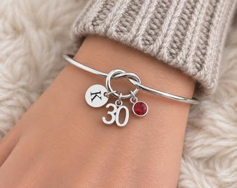 May include: A silver-toned bangle bracelet featuring a central knot design. Three charms hang from the knot: a circular disc engraved with the letter K, the number 30 in a bold font, and a small, round, faceted red crystal set in silver.