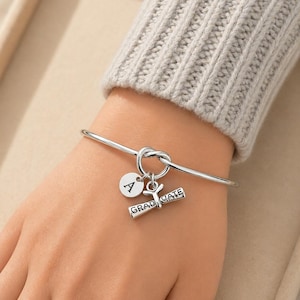 May include: A silver-colored bangle bracelet with a knot design. The bracelet features two charms: a round charm with the letter "A" and a diploma charm with the word "GRADUATE".