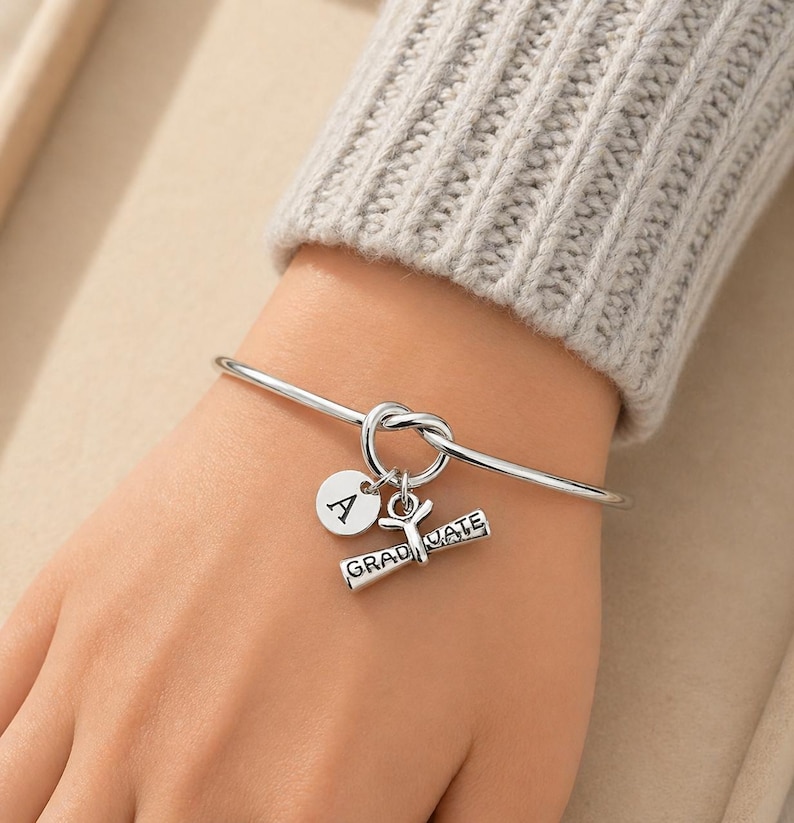 May include: A silver-colored bangle bracelet with a knot design. The bracelet features two charms: a round charm with the letter "A" and a diploma charm with the word "GRADUATE".