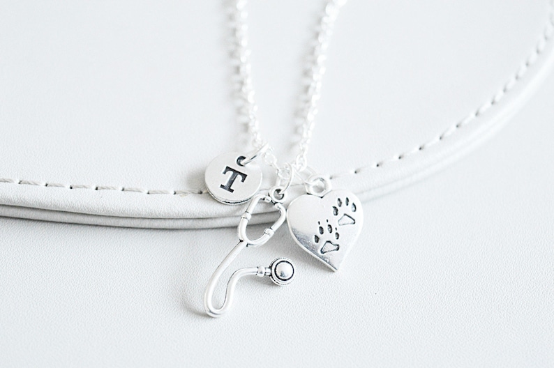 Peut inclure: Un collier argent&eacute; avec trois breloques : un st&eacute;thoscope, un c&oelig;ur avec des empreintes de pattes et un cercle avec la lettre "T". Le collier est pr&eacute;sent&eacute; sur une surface blanche, &eacute;voquant un th&egrave;me v&eacute;t&eacute;rinaire ou m&eacute;dical.