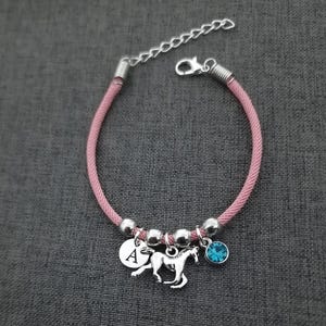 K&ouml;nnte beinhalten: Rosa geflochtenes Armband mit einem silbernen Pferdeanh&auml;nger, einem silbernen Anh&auml;nger mit dem Buchstaben A und einem hellblauen Kristallanh&auml;nger.