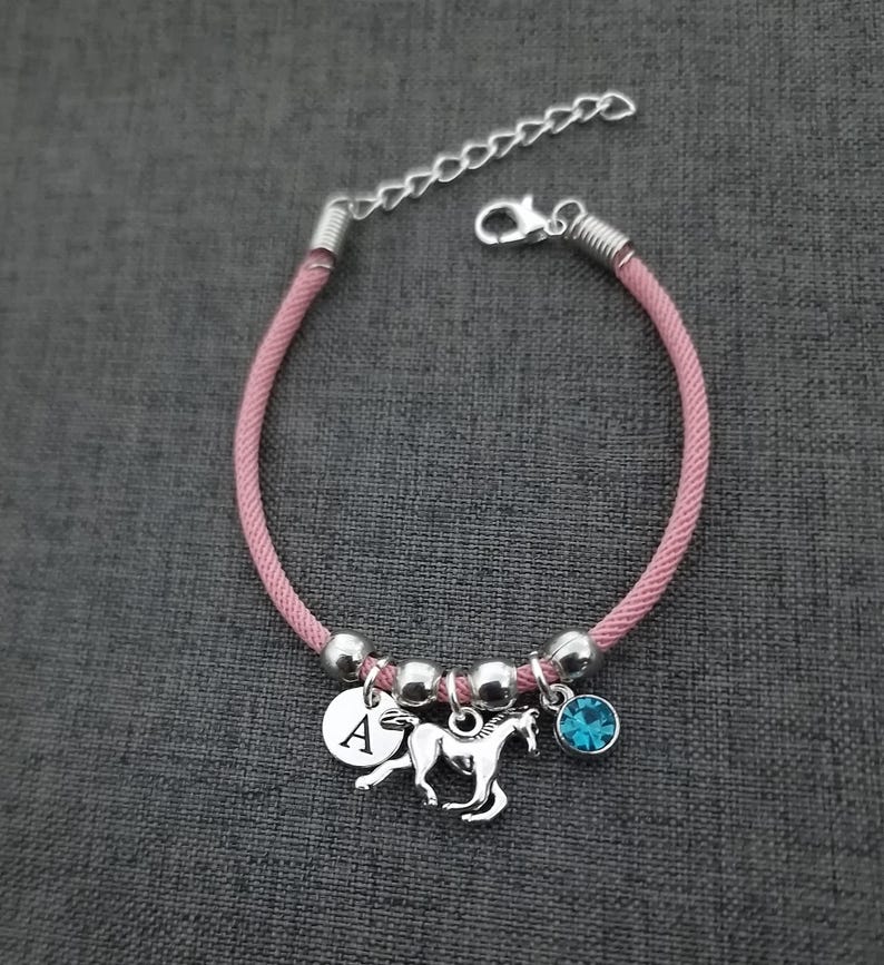 K&ouml;nnte beinhalten: Rosa geflochtenes Armband mit einem silbernen Pferdeanh&auml;nger, einem silbernen Anh&auml;nger mit dem Buchstaben A und einem hellblauen Kristallanh&auml;nger.