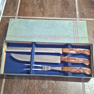 May include: A three-piece cutlery set in a green box with a blue interior. The set includes a serrated knife, a carving knife, and a carving fork, all featuring brown marbled handles and stainless steel blades marked Sheffield Stainless Blade.