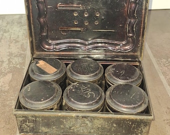 May include: An antique metal box with a hinged lid, containing six small, round, black containers. The box and containers show signs of age and wear, with a distressed finish. The lid has an ornate design.