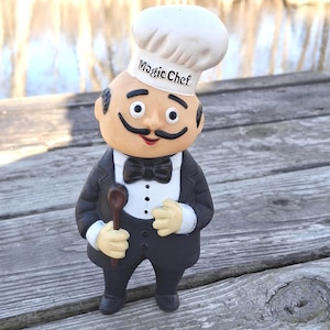 May include: A figurine of a chef wearing a white hat with the text "Magic Chef", a black bow tie, and a black suit. The chef is holding a wooden spoon. The figurine has a large mustache and is standing on a wooden surface.