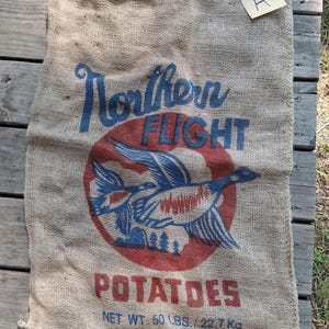 May include: A tan burlap sack featuring the blue text Northern Flight above a red circle with a blue duck illustration. Below the graphic, the red text POTATOES appears, followed by blue text stating NET WT 50 LBS / 22.7 KG PRODUCE OF U.S.A.