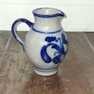 May include: A small, off-white ceramic pitcher with a dark blue handle and spout. The pitcher is decorated with blue floral designs. The base and rim are also trimmed in blue. The pitcher is likely used for serving liquids.