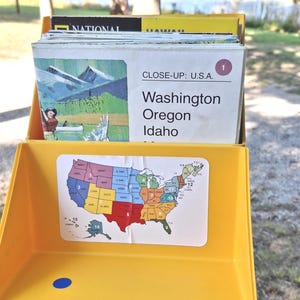 May include: A yellow plastic storage box containing a stack of National Geographic educational cards. The front of the box features a colorful map of the U.S.A. The top card is titled CLOSE-UP: U.S.A. and lists Washington, Oregon, and Idaho with an illustration.