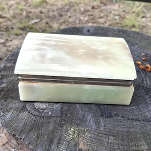 May include: A rectangular box crafted from polished light green and white marbled stone. The lid features a smooth, translucent surface with natural veining, while a thin silver-toned metal band separates the lid from the base of the container.