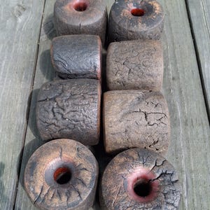 May include: A collection of eight cylindrical wooden objects with a dark, charred, and cracked exterior texture. Each piece features a central circular opening, revealing a smooth, reddish-brown interior surface within the core of the wood.