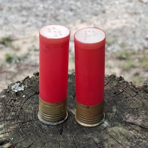 May include: Two red and brass shotgun shell salt and pepper shakers. The shakers have a white top with multiple holes for dispensing. They are cylindrical in shape and sit on a weathered wooden surface.