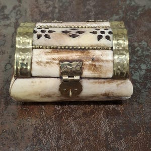 May include: A small, rectangular jewelry box with a hinged lid. The box is made of a light-colored material, possibly bone, with brass accents. The lid features a decorative cutout pattern and a brass clasp. The box is on a brown surface.