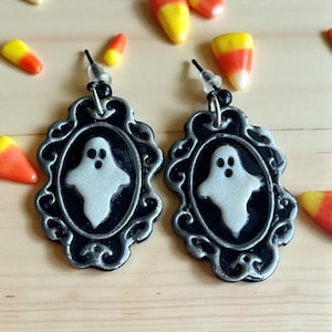Op de afbeelding: Een paar Halloween-oorbellen. Elke oorbel heeft een zilveren spookontwerp op een zwarte ovaal, omlijst door een decoratieve zilveren rand. Snoepmaïssnoepjes zijn verspreid rond de oorbellen.