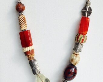 May include: A brown leather cord necklace with a silver pendant featuring a starburst design. The necklace is adorned with various beads, including red, orange, brown, and white.