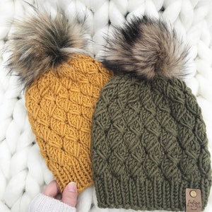May include: Two knitted hats with faux fur pom poms. One hat is mustard yellow and the other is olive green. Both hats have a diamond knit pattern. The green hat has a brown leather tag with the text "Indigo Mint Co."
