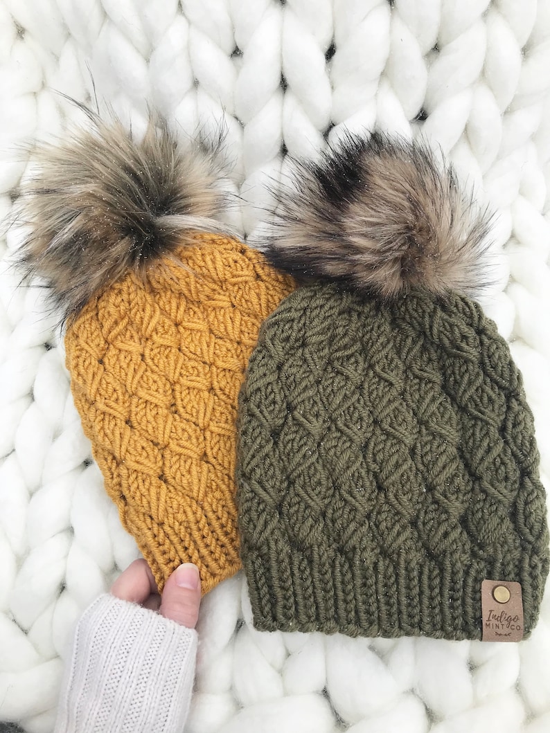 May include: Two knitted hats with faux fur pom poms. One hat is mustard yellow and the other is olive green. Both hats have a diamond knit pattern. The green hat has a brown leather tag with the text "Indigo Mint Co."