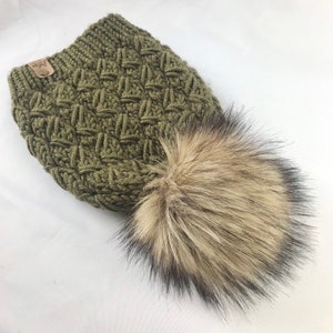 May include: A green knitted beanie with a faux fur pom pom. The beanie has a textured pattern and a small brown leather tag with the word "Cozy" on it.