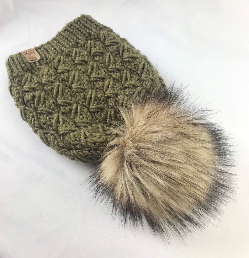 May include: A green knitted beanie with a faux fur pom pom. The beanie has a textured pattern and a small brown leather tag with the word "Cozy" on it.