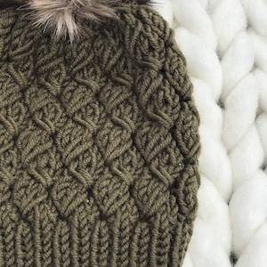 May include: A green knitted beanie with a fluffy brown and black pom pom. The beanie has a cable knit pattern.