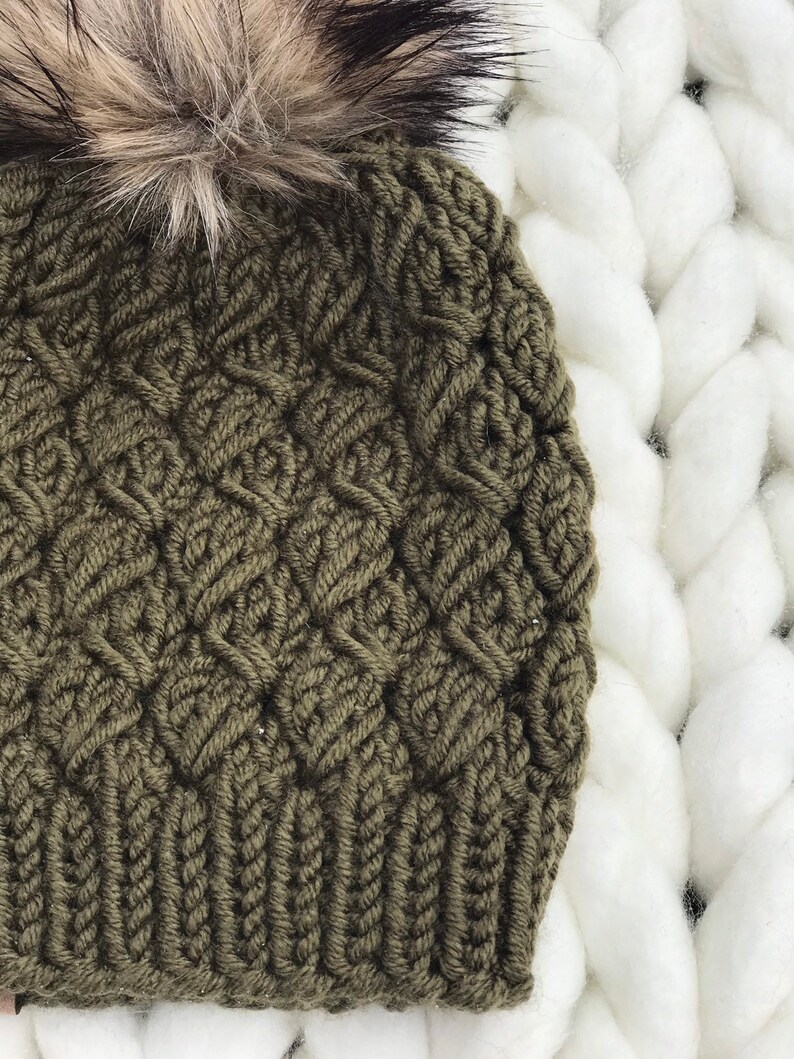 May include: A green knitted beanie with a fluffy brown and black pom pom. The beanie has a cable knit pattern.