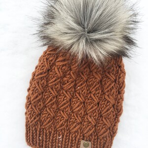 May include: A brown knitted beanie with a large white and gray faux fur pom pom. The beanie has a woven pattern and a brown leather tag that says "Indigo Mint Co."