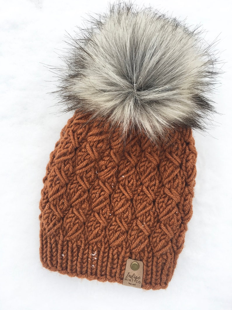 May include: A brown knitted beanie with a large white and gray faux fur pom pom. The beanie has a woven pattern and a brown leather tag that says "Indigo Mint Co."