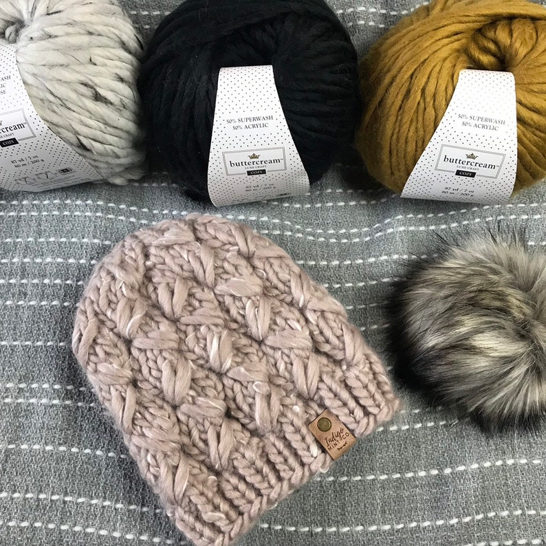 May include: A pink knitted beanie with a cable knit pattern and a brown leather tag that says "Indigo Knits". The beanie is sitting on a gray and white striped blanket with two balls of yarn in the background.