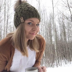 May include: A woman wearing a green knit hat with a pom pom and a brown cardigan holds a white ceramic mug with the text "GET AFTER IT". The mug is filled with a brown liquid. The woman is standing in a snowy forest.