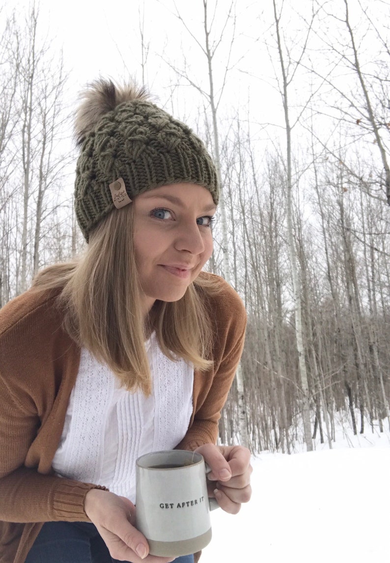 May include: A woman wearing a green knit hat with a pom pom and a brown cardigan holds a white ceramic mug with the text "GET AFTER IT". The mug is filled with a brown liquid. The woman is standing in a snowy forest.