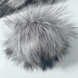 May include: A fluffy gray and black faux fur pom pom. The pom pom is round and has a soft, fuzzy texture.