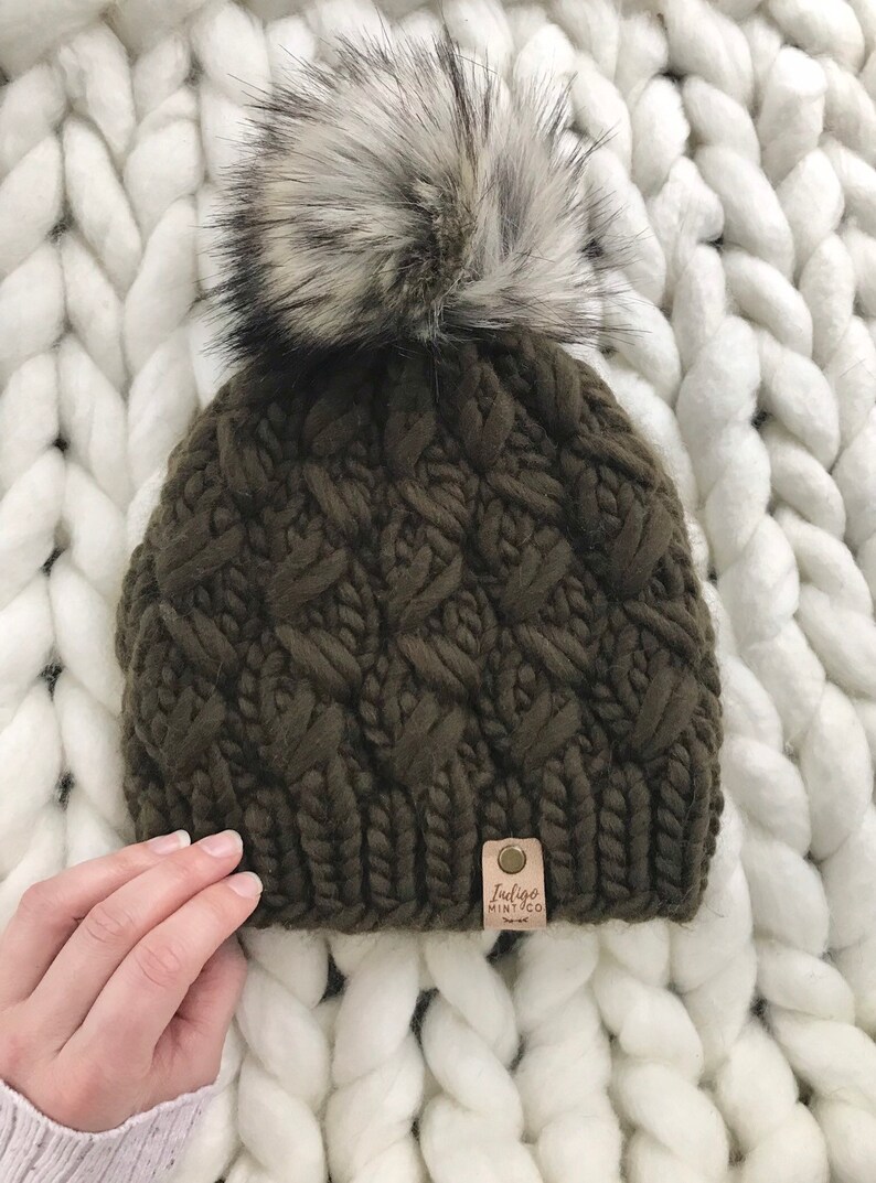 May include: A dark green knitted beanie with a large white and black faux fur pom pom. The beanie has a cable knit pattern and a small brown leather tag with the text "Indigo Mint Co."