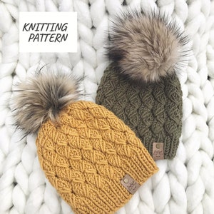May include: Two knitted hats with faux fur pom poms. The hats are in a cable knit pattern. One hat is mustard yellow and the other is olive green. Both hats have a brown leather tag with the text "Indigo Knit Co."