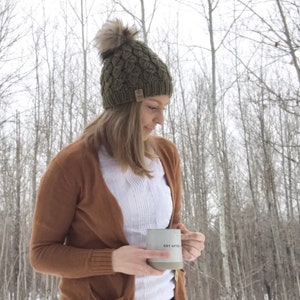 May include: A woman wearing a brown cardigan, white shirt, and blue jeans is holding a gray ceramic mug with the text "Get After It" in black. She is wearing a green knit beanie with a faux fur pom pom. The background is a snowy forest.