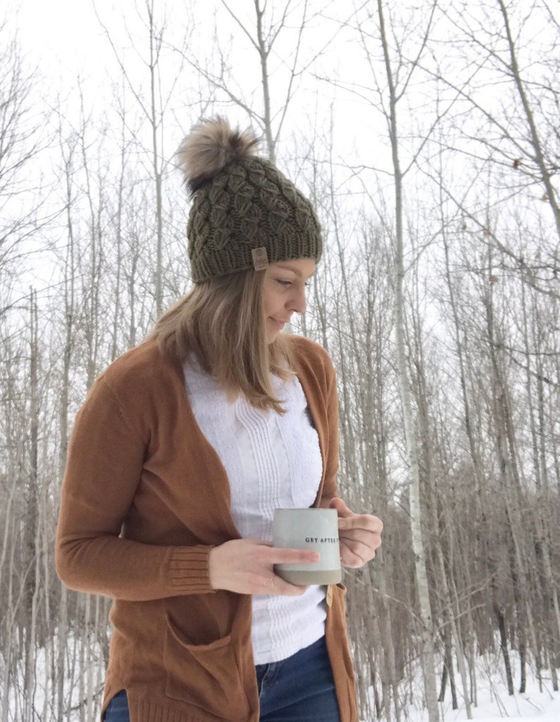 May include: A woman wearing a brown cardigan, white shirt, and blue jeans is holding a gray ceramic mug with the text "Get After It" in black. She is wearing a green knit beanie with a faux fur pom pom. The background is a snowy forest.