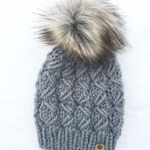 May include: Gray knitted beanie with a large, fluffy white and gray pom pom. The beanie has a cable knit pattern and a brown leather tag with the text "Indigo Mint Co."