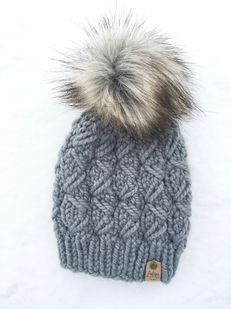 May include: Gray knitted beanie with a large, fluffy white and gray pom pom. The beanie has a cable knit pattern and a brown leather tag with the text "Indigo Mint Co."