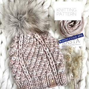 May include: A pink and white knitted beanie with a fluffy pom pom on top. The beanie is made with a chunky yarn and has a textured stitch pattern. The yarn label reads "Rasta 969 Whole Grain".