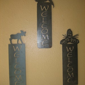 May include: Three metal welcome signs with animal silhouettes. The signs are rectangular with the word "WELCOME" cut out. One sign has a moose, another a bird, and the third a pinecone.